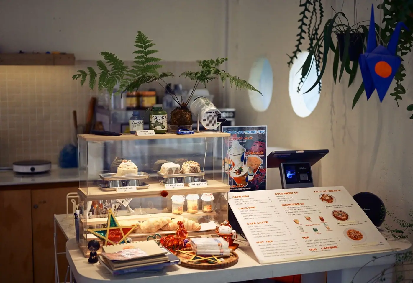Cozy coffee shop interior with a pastry display, ferns, and a menu on the counter.