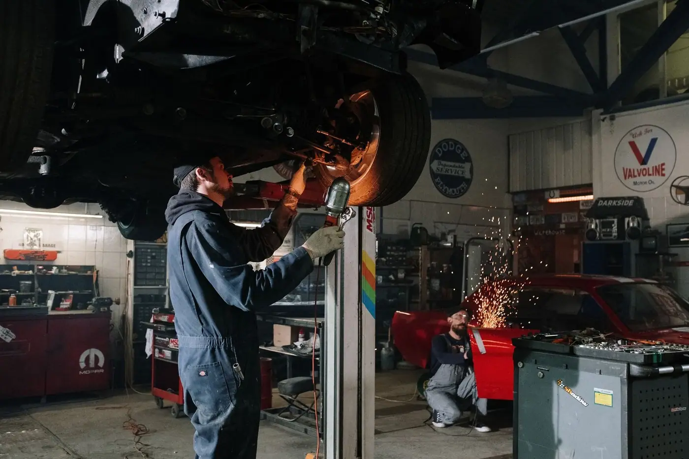 Two automotive technicians repairing a car in a busy workshop with sparks flying.