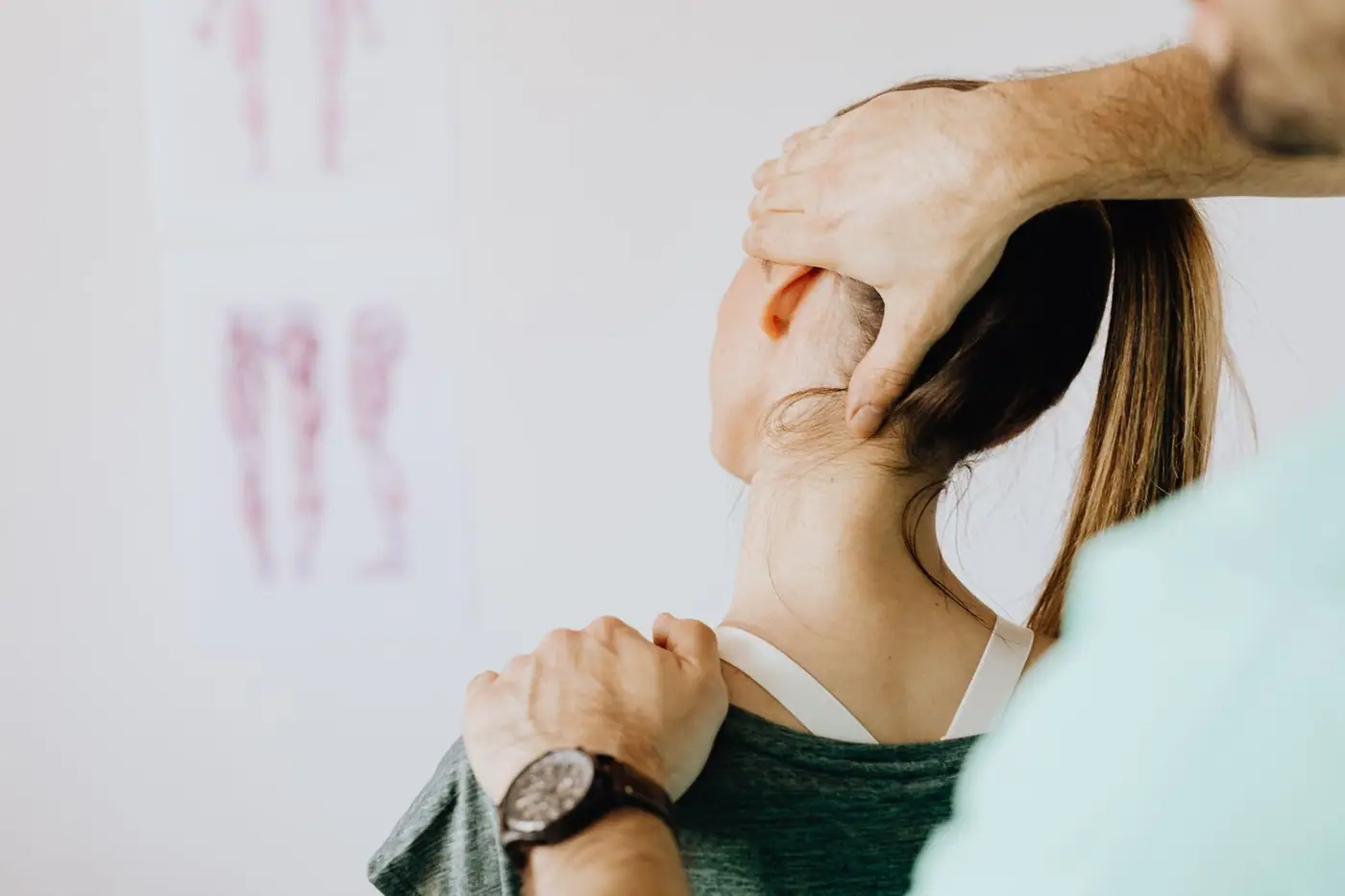 Back view of crop faceless bearded orthopedist in wristwatch checking up neck of anonymous lady while standing in front of wall with paper d