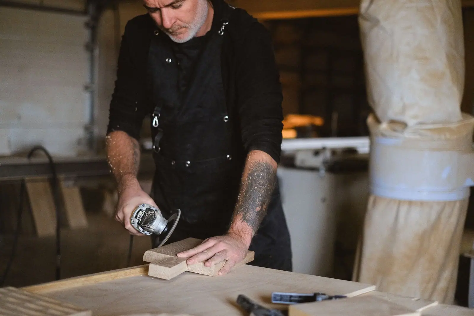 Focused male master in apron with angle grinder working with wooden board while standing at table with special instruments in workshop