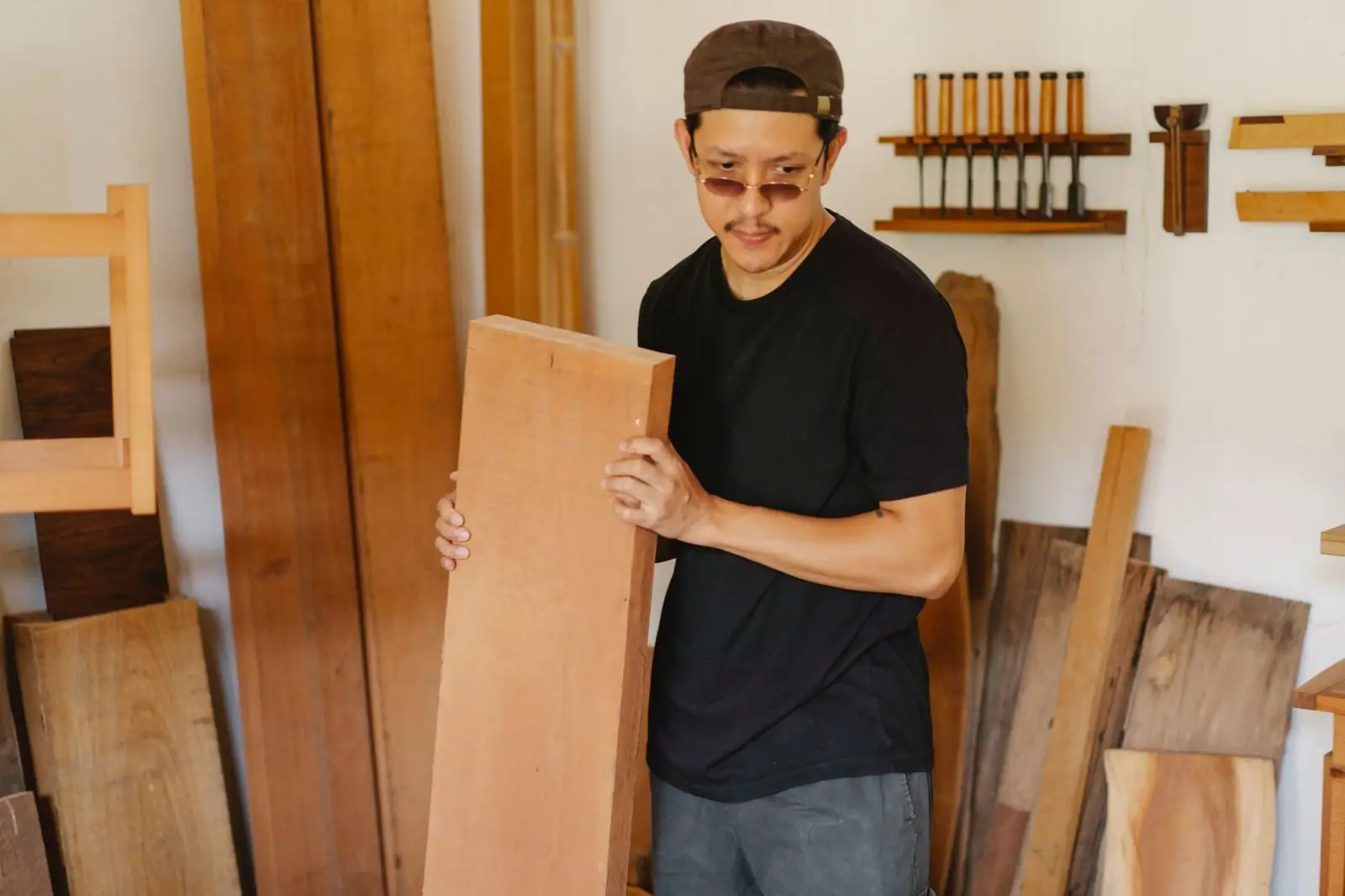 Serious ethnic male in casual outfit pondering with timber plank in arms in workshop