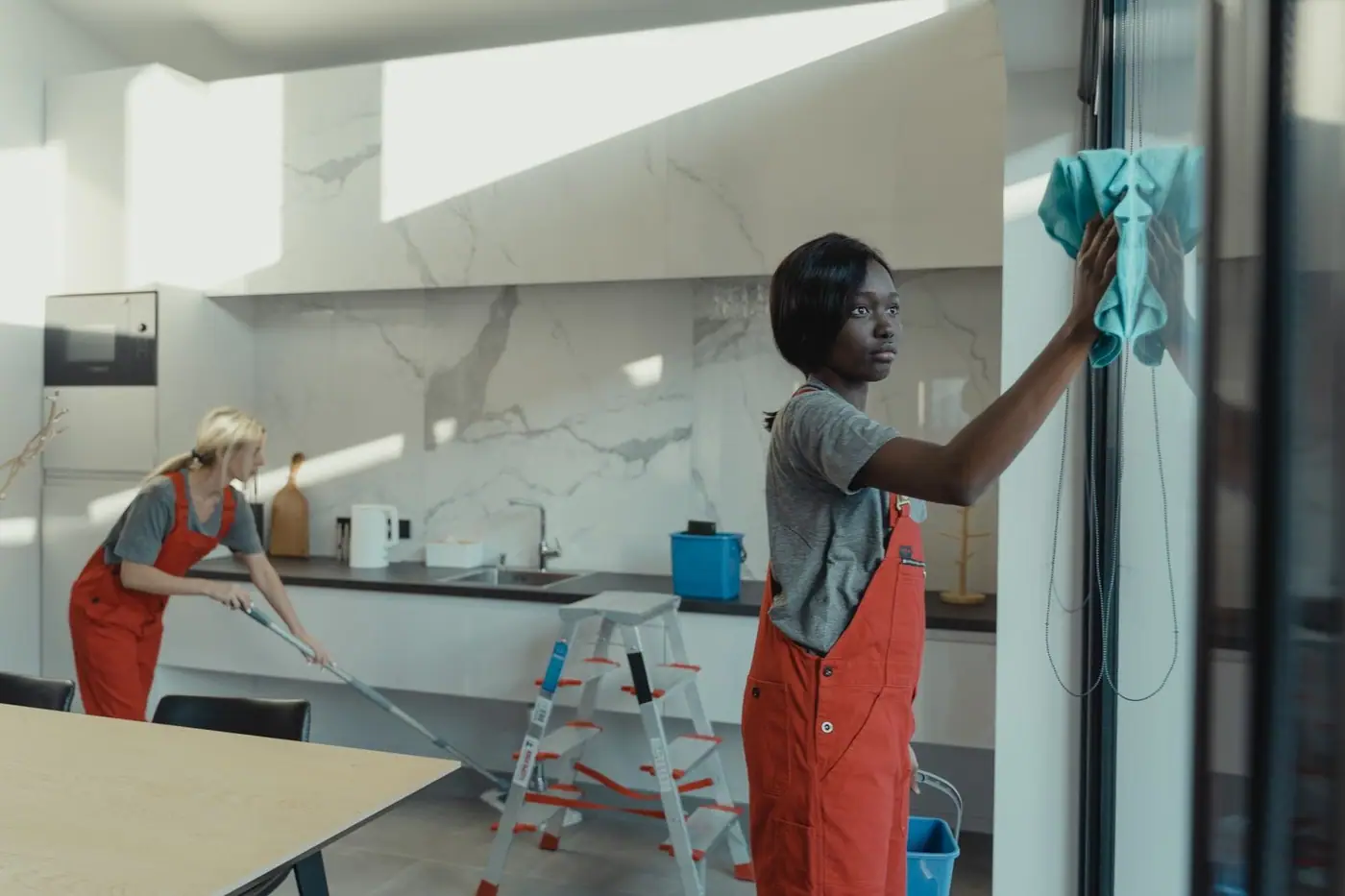 Two women cleaning a modern kitchen, wiping glass surfaces and mopping the floor