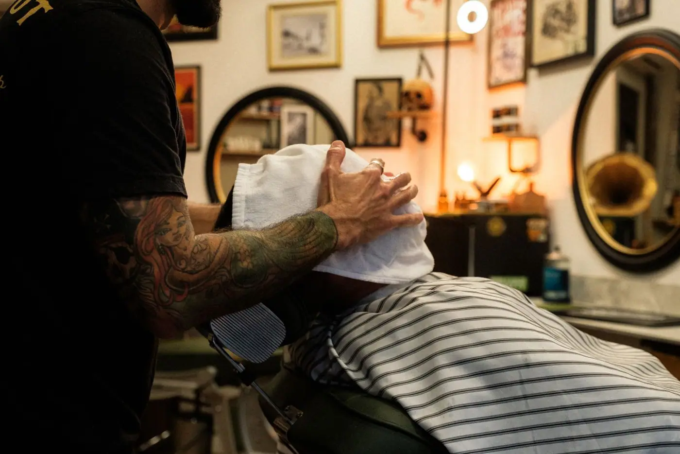 A barber with tattoo sleeves applies a hot towel in a vintage-style barbershop setting.